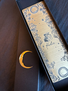 A gold bookmark with black decals of 2 moons, a stack of books, flowers and personalised with the name Kadie inside a gift box.