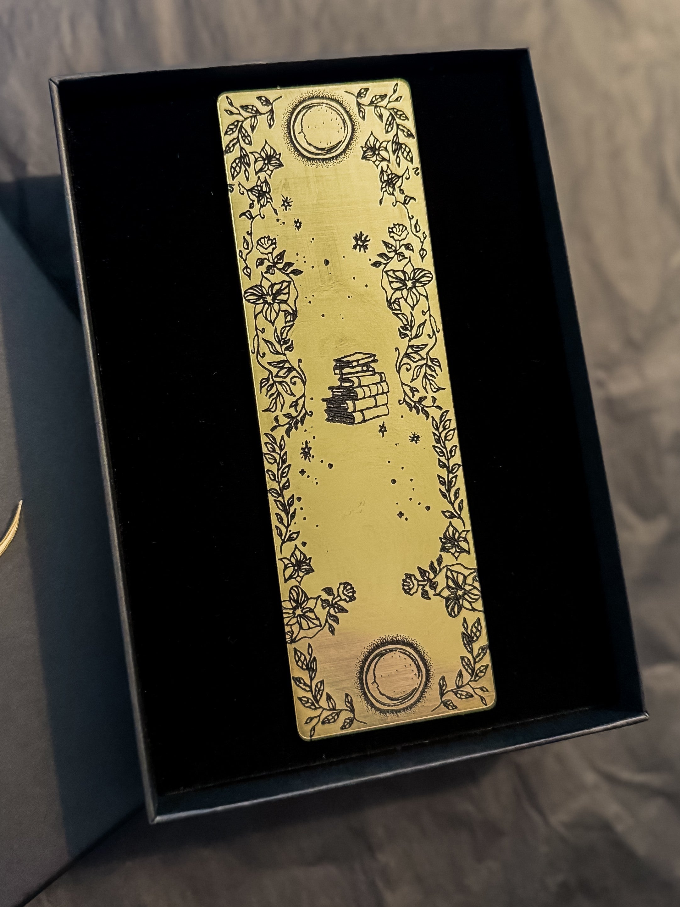 A gold bookmark with black illustrations of a book, two moons & flowers.