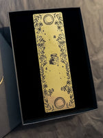 Load image into Gallery viewer, A gold bookmark with black illustrations of a book, two moons &amp; flowers.
