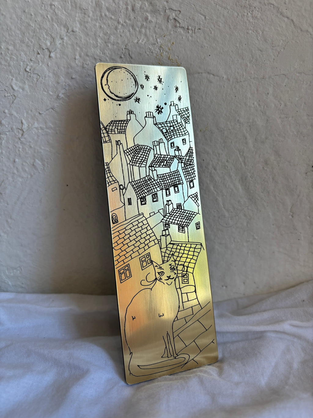 Gold bookmark with black image of cat, houses & moon.