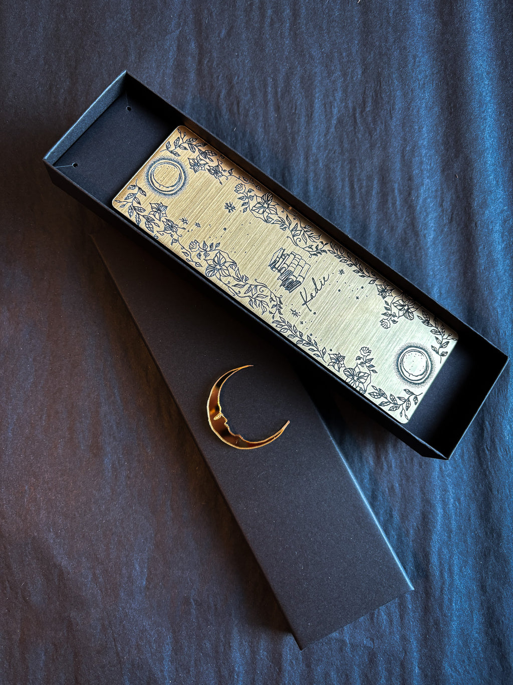 A gold bookmark with black decals of 2 moons, a stack of books, flowers and personalised with the name Kadie.