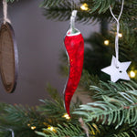 Load image into Gallery viewer, A long chilli pepper shaped ornament hanging in a christmas tree next to a star shaped bauble.
