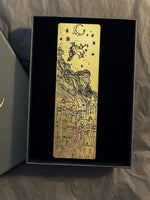 Load image into Gallery viewer, Gold bookmark with black images of witch on broomstick above buildings.
