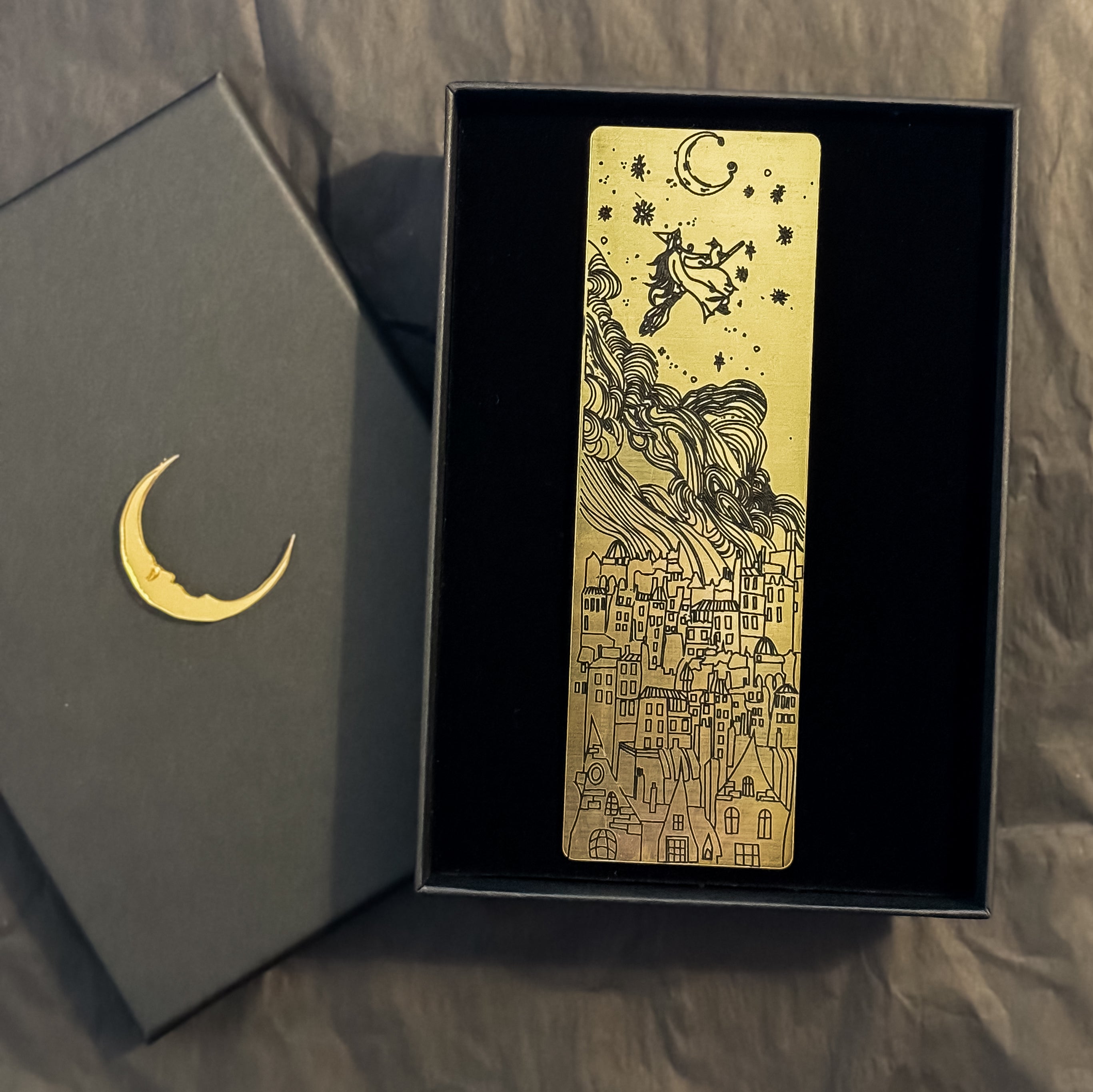 Gold bookmark with black images of witch on broomstick above buildings, in a gift box.