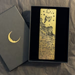 Load image into Gallery viewer, Gold bookmark with black images of witch on broomstick above buildings, in a gift box.
