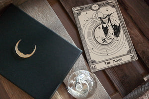 A decorative tarot card with a bat hanging under the moon next to gift box.