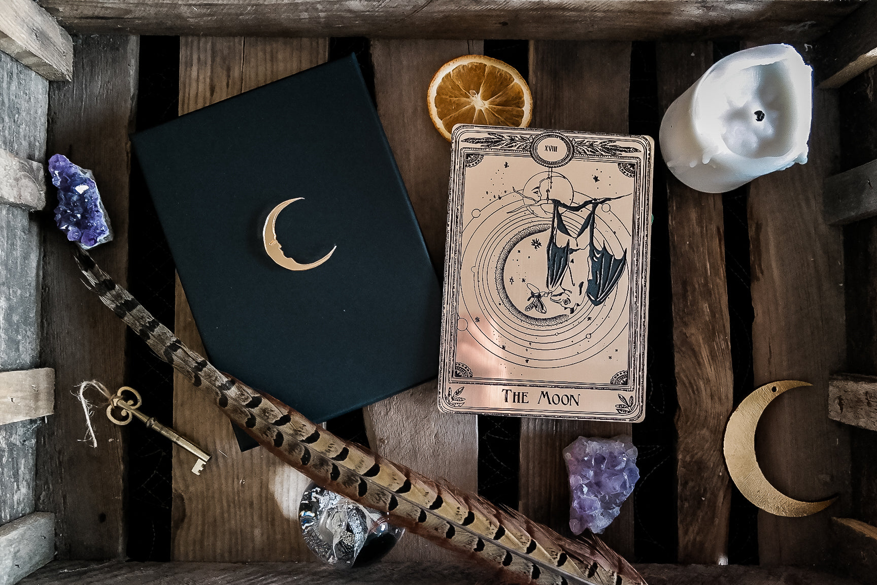 A decorative tarot card with a bat hanging under the moon next to gift box, candle, crystals and feather.