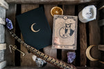 Load image into Gallery viewer, A decorative tarot card with a bat hanging under the moon next to gift box, candle, crystals and feather.

