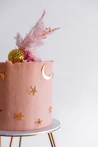 A pink birthday cake with gold cake charms in shape of crescent moon and stars with golden disco ball topper