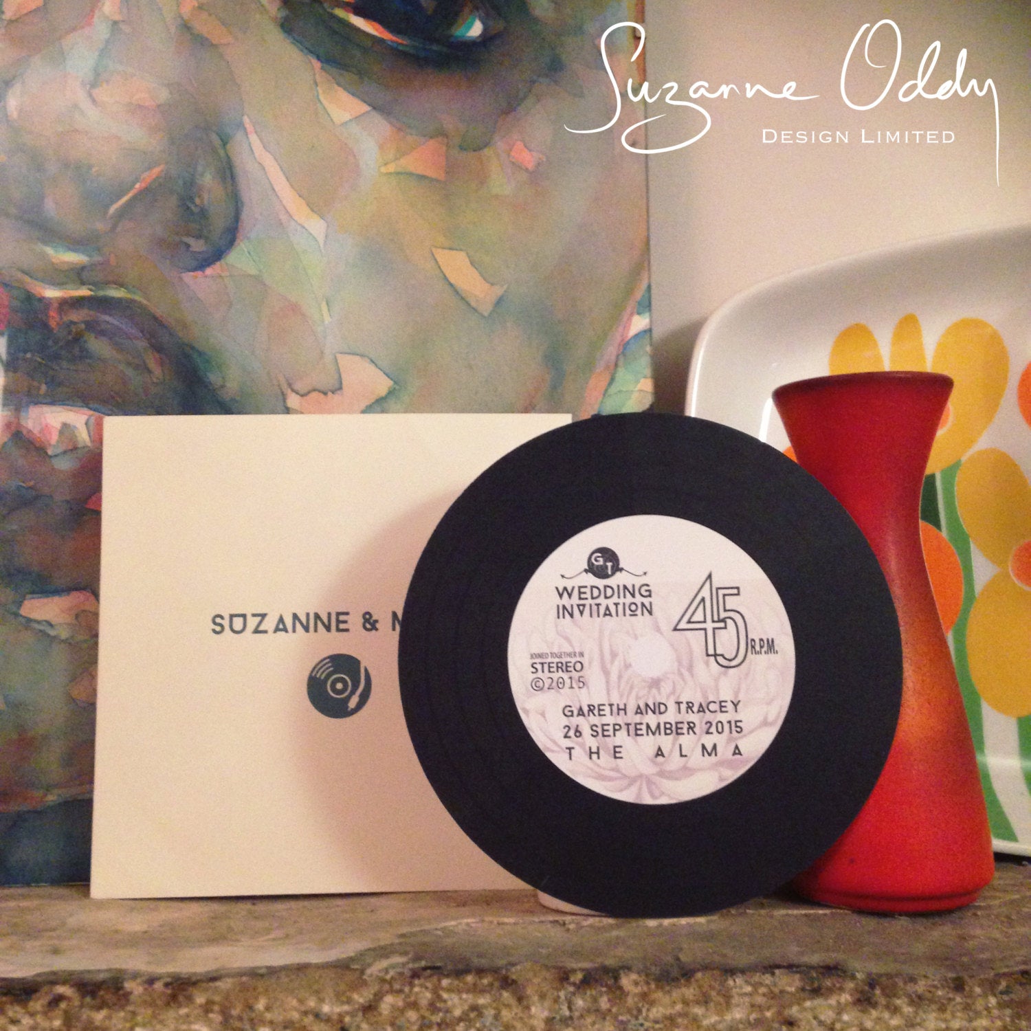 A wedding invitation shaped like a record next to red vase.
