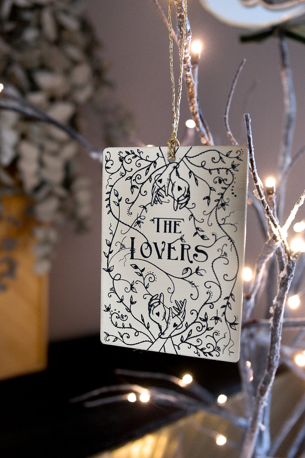 A tarot card ornament for hanging with the words 'The Lovers' etched in by hand with vine leaves and hands intertwined. 